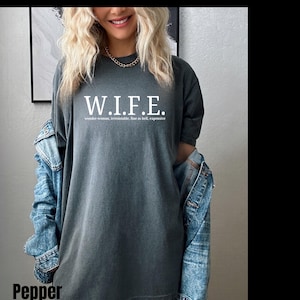 Pu&ograve; includere: Una t-shirt a maniche corte color grigio antracite con la scritta bianca W.I.F.E. e la definizione wonder-woman, irresistable, fine as hell, expensive stampata sotto. La t-shirt &egrave; indossata con una giacca di jeans blu sulle spalle.
