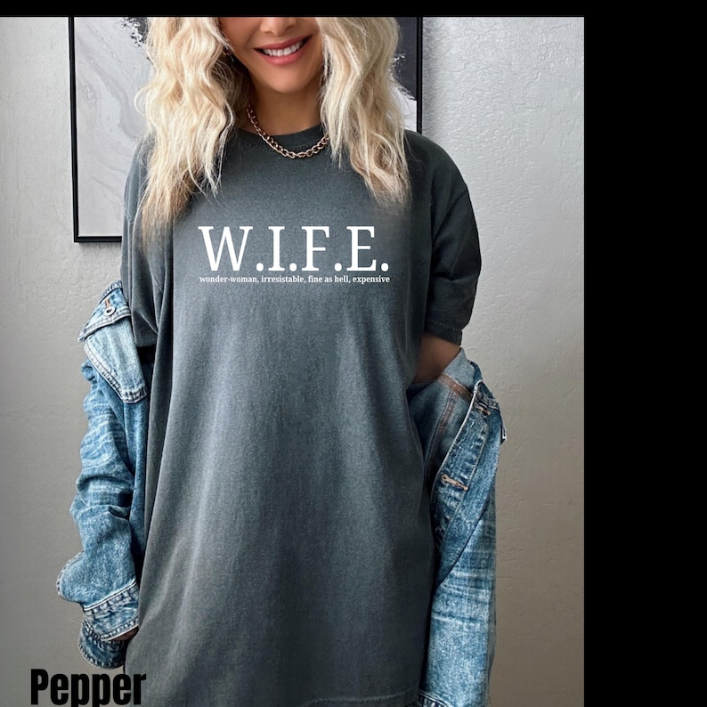 Pu&ograve; includere: Una t-shirt a maniche corte color grigio antracite con la scritta bianca W.I.F.E. e la definizione wonder-woman, irresistable, fine as hell, expensive stampata sotto. La t-shirt &egrave; indossata con una giacca di jeans blu sulle spalle.