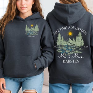 May include: Dark grey hoodie with a front pocket and hood. The front has a nature scene with trees, a river, and a sun, with the text "EST 2026". The back displays "LIFETIME ADVENTURE" with a similar design and the text "Mr. & Mrs. BARSTEN".