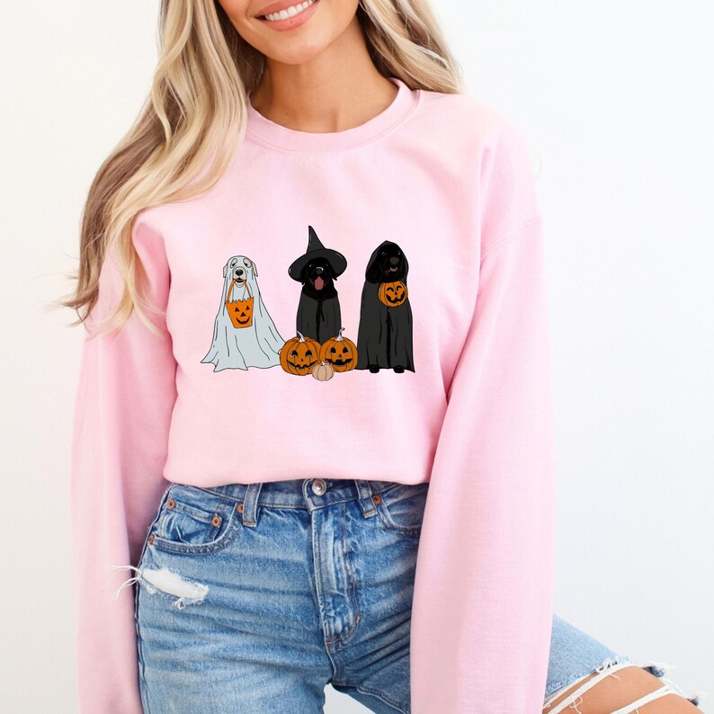 May include: A light pink sweatshirt featuring a Halloween-themed graphic. The graphic depicts three dogs dressed in Halloween costumes: a ghost, a witch, and a grim reaper, each holding a pumpkin-shaped treat bag. The dogs are positioned in front of carved pumpkins.