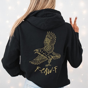 Op de afbeelding: Zwarte hoodie met een gouden illustratie van een raaf met uitgespreide vleugels. De tekst "F-CAW-F" staat eronder. De hoodie heeft een capuchon en lange mouwen. De achtergrond is effen wit.
