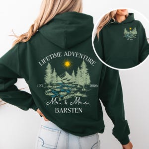 Może przedstawiać: Ciemnozielona bluza z kapturem z krajobrazem przedstawiającym drzewa, góry, słońce i napis "LIFETIME ADVENTURE". Poniżej słowa "Mr. & Mrs. BARSTEN" i rok "2026".