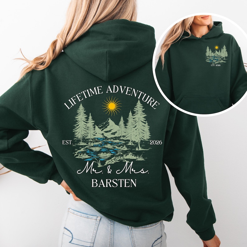 May include: Dark green hoodie with a landscape design featuring trees, mountains, a sun, and the text "LIFETIME ADVENTURE". Below the graphic are the words "Mr. & Mrs. BARSTEN" and the year "2026".