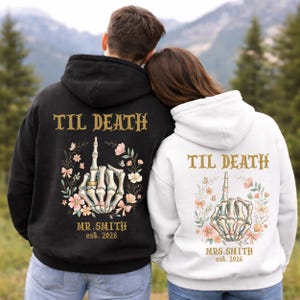 May include: Black and white hoodies with the text "TIL DEATH" in gold lettering. The design features a skeleton hand with a ring, surrounded by flowers and bows. The text "MR. SMITH est. 2026" and "MRS. SMITH est. 2026" are also included.