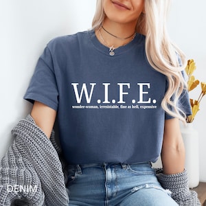 Pu&ograve; includere: Una t-shirt blu a maniche corte con il testo bianco W.I.F.E. in un grande carattere serif, con il testo bianco pi&ugrave; piccolo wonder-woman, irresistible, fine as hell, expensive stampato direttamente sotto l'acronimo sul petto.