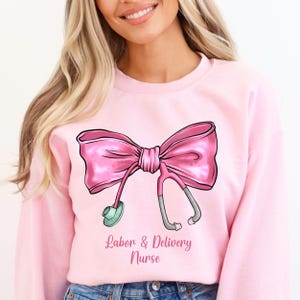 Op de afbeelding: Roze sweatshirt met een roze strik en stethoscoop ontwerp. De tekst "Labor & Delivery Nurse" is onder het ontwerp gedrukt.