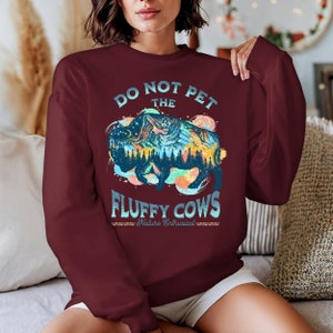 Pu&ograve; includere: Una felpa color bordeaux con una grafica di un bisonte con un paesaggio montano al suo interno. Il testo "Do Not Pet The Fluffy Cows" &egrave; stampato sulla felpa. Il testo "Nature Enthusiast" &egrave; stampato sotto la grafica.