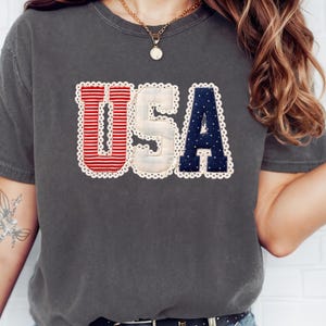 Pode incluir: T-shirt cinzenta escura com a palavra "USA" em riscas vermelhas e brancas, branco e azul marinho com detalhes brancos em forma de estrela. As letras são delineadas com uma borda de renda branca. A t-shirt é de estilo casual.
