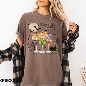 May include: A brown t-shirt with a graphic of three witches' brew cocktails, a black cat, and the text "Witches Brew Guest Check Pick Your Poison".