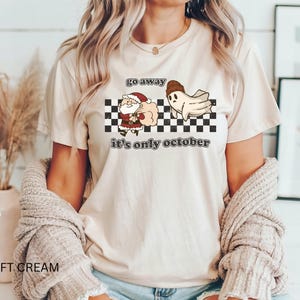 May include: A cream-colored t-shirt with a graphic of a ghost chasing Santa Claus away with the text "Go away it's only October".