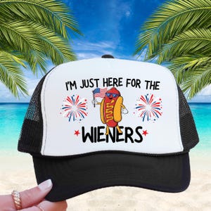 Puede incluir: Una gorra de camionero blanca y negra con una bandera estadounidense roja, blanca y azul y un perro caliente de dibujos animados sosteniendo la bandera. El texto en la gorra dice "I'm just here for the wieners".