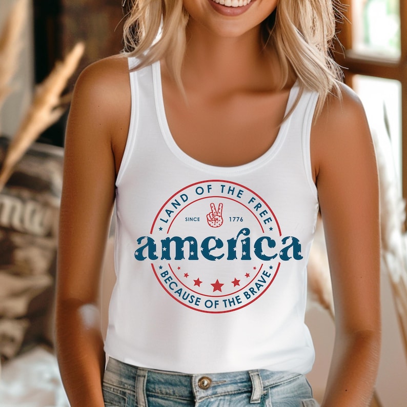 Pode incluir: Top branco com um design patri&oacute;tico. O design apresenta a palavra "america" em azul, circundada por um c&iacute;rculo vermelho com o texto "Land of the Free" e "Because of the Brave", juntamente com estrelas e um sinal de paz.