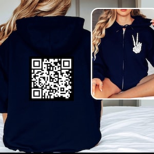May include: A navy blue zippered hoodie with a white QR code on the back. The hoodie has a hood and a zipper closure. The model is wearing the hoodie and has long blonde hair.