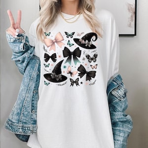 Pode incluir: Camiseta branca de manga comprida com uma estampa gr&aacute;fica de chap&eacute;us de bruxa pretos, la&ccedil;os rosa e pretos e borboletas. O gr&aacute;fico est&aacute; centralizado na camiseta.