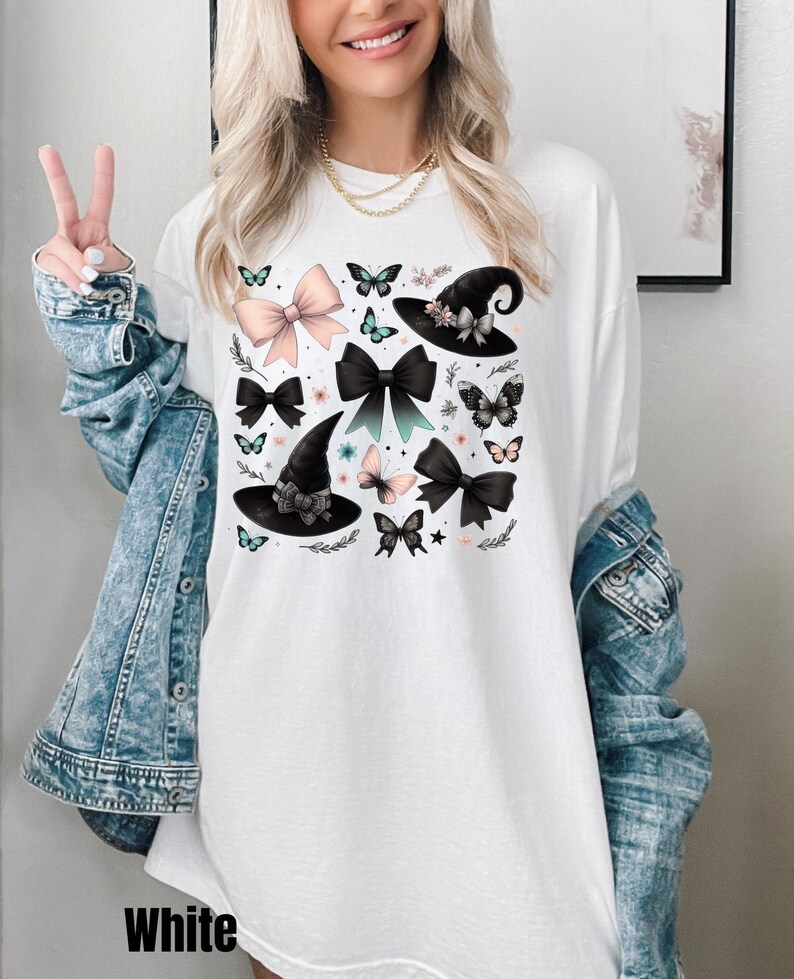 Pode incluir: Camiseta branca de manga comprida com uma estampa gr&aacute;fica de chap&eacute;us de bruxa pretos, la&ccedil;os rosa e pretos e borboletas. O gr&aacute;fico est&aacute; centralizado na camiseta.