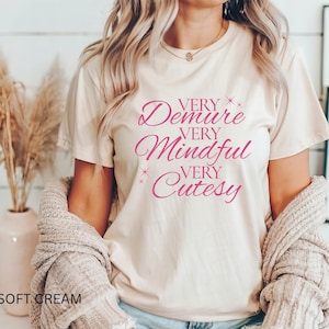 以下が含まれることがあります： ピンクの文字で「Very Demure Very Mindful Very Cutesy」とプリントされた白いTシャツです。