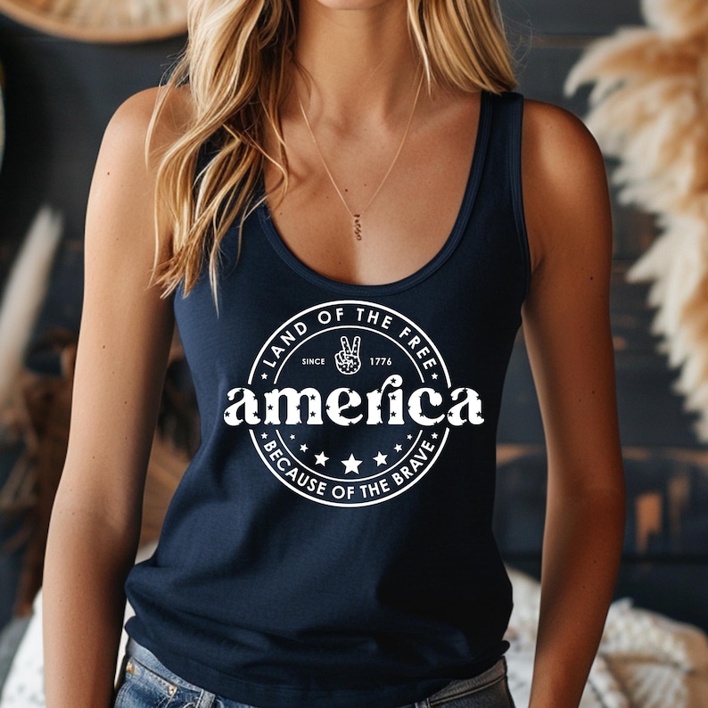 Pode incluir: Top sem mangas azul marinho com um gr&aacute;fico circular branco que diz "Land of the Free Since 1776 America Because of the Brave" com um s&iacute;mbolo de paz. O top sem mangas &eacute; sem mangas e tem decote redondo.