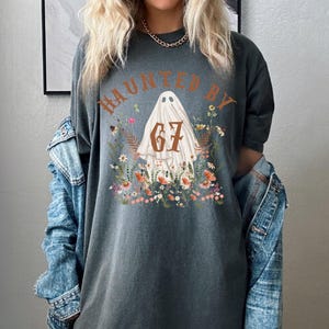 Puede incluir: Camiseta gris oscuro con el texto "HAUNTED BY 67" sobre una ilustraci&oacute;n de fantasma rodeada de flores. La camiseta es de corte holgado y tiene mangas cortas. La palabra "Pepper" est&aacute; impresa en la parte inferior izquierda.