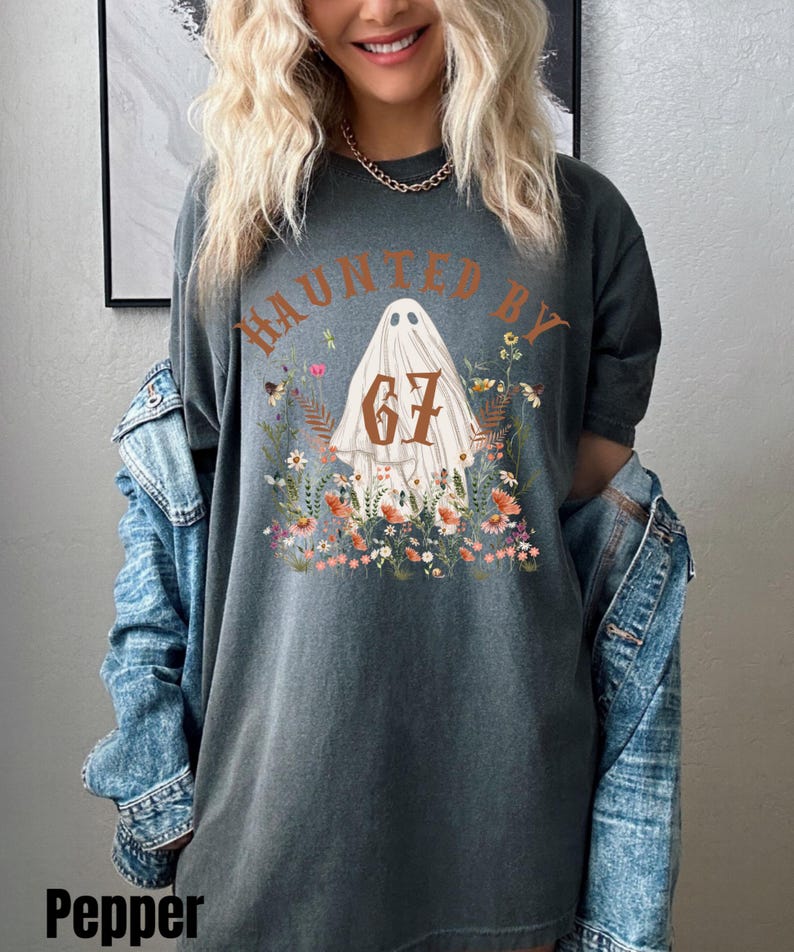 Puede incluir: Camiseta gris oscuro con el texto "HAUNTED BY 67" sobre una ilustraci&oacute;n de fantasma rodeada de flores. La camiseta es de corte holgado y tiene mangas cortas. La palabra "Pepper" est&aacute; impresa en la parte inferior izquierda.