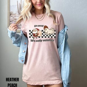 May include: A pink t-shirt with a black and white checkered background. The shirt has a graphic of a ghost chasing Santa Claus away with the text "go away it's only october".
