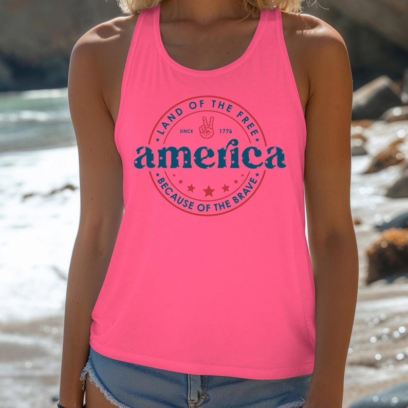 Pode incluir: Regata rosa brilhante com um gr&aacute;fico redondo que diz "Land of the Free" e "America" em azul, com um sinal de paz e estrelas. O texto "Because of the Brave" tamb&eacute;m est&aacute; inclu&iacute;do. A regata &eacute; sem mangas.