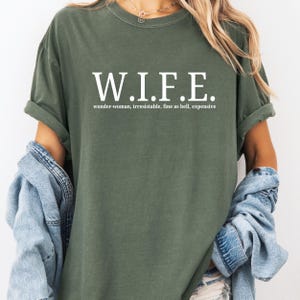 Pu&ograve; includere: Una t-shirt a maniche corte color verde oliva con il testo bianco W.I.F.E. stampato in un carattere serif in grassetto. Sotto l'acronimo, il testo recita wonder-woman, irresistible, fine as hell, expensive in un carattere sans-serif pi&ugrave; piccolo.