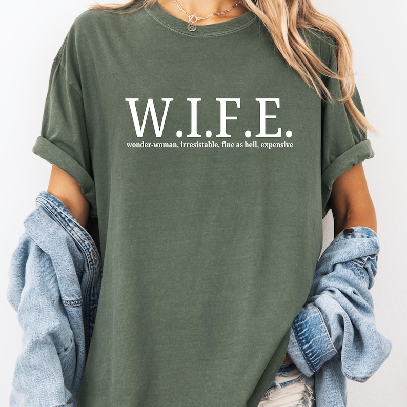 Pu&ograve; includere: Una t-shirt a maniche corte color verde oliva con il testo bianco W.I.F.E. stampato in un carattere serif in grassetto. Sotto l'acronimo, il testo recita wonder-woman, irresistible, fine as hell, expensive in un carattere sans-serif pi&ugrave; piccolo.