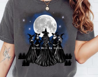 May include: A dark gray t-shirt with a graphic of three witches standing in a circle under a full moon. The text "Girls Will Be Girls." is printed below the witches.