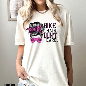Puede incluir: Una camiseta blanca con un gr&aacute;fico de una mujer con un mo&ntilde;o desordenado, gafas de sol y una diadema de leopardo rosa. El texto "Bike Hair. Don't Care." est&aacute; impreso en la camiseta.