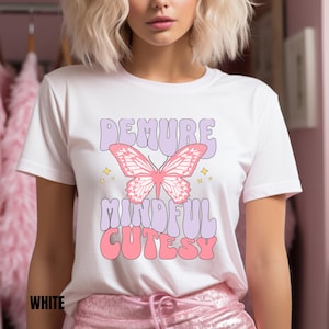 以下が含まれることがあります： レトロなフォントで「DEMURE MINDFUL CUTESY」というテキストとピンクの蝶のグラフィックが描かれた白いTシャツです。