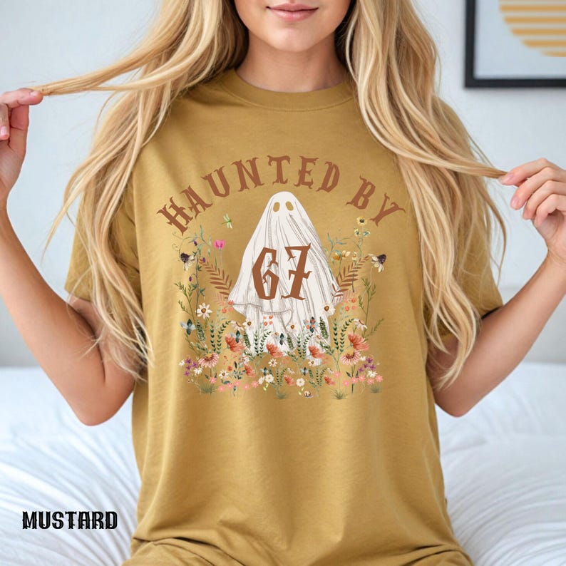 Puede incluir: Camiseta color mostaza con el texto "HAUNTED BY 67" sobre una ilustraci&oacute;n de fantasma rodeada de flores. La camiseta tiene un corte holgado, perfecta para Halloween o la temporada de miedo.