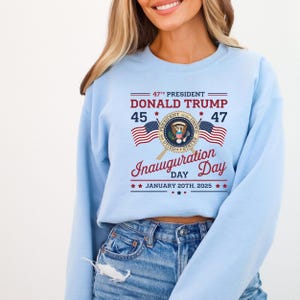 以下が含まれることがあります： 薄い青色のスウェットシャツに、大統領の紋章、アメリカ国旗、そして「47th President Donald Trump 45 47 Inauguration Day January 20th, 2025」というテキストがプリントされています。