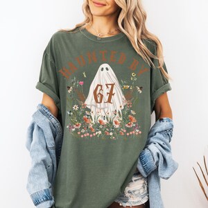 Puede incluir: Camiseta verde oliva con el texto "HAUNTED BY 67" en marr&oacute;n. El dise&ntilde;o presenta un fantasma blanco rodeado de flores coloridas. La camiseta tiene mangas cortas y un corte holgado, ideal para Halloween.