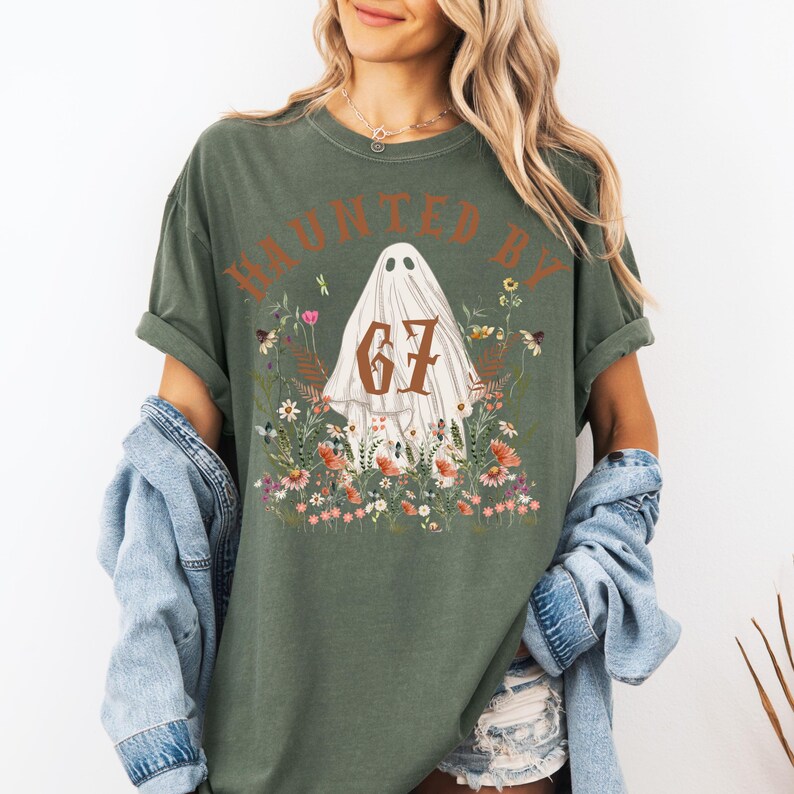 Puede incluir: Camiseta verde oliva con el texto "HAUNTED BY 67" en marr&oacute;n. El dise&ntilde;o presenta un fantasma blanco rodeado de flores coloridas. La camiseta tiene mangas cortas y un corte holgado, ideal para Halloween.