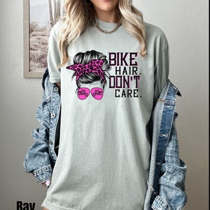 Puede incluir: Una camiseta verde claro con un gr&aacute;fico de una mujer con un mo&ntilde;o desordenado, una bandana rosa y gafas de sol. Las gafas de sol tienen un gr&aacute;fico de una motocicleta. El texto en la camiseta dice "Bike Hair Don't Care."
