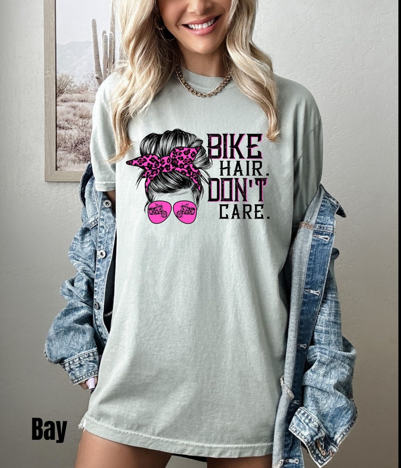 Puede incluir: Una camiseta verde claro con un gr&aacute;fico de una mujer con un mo&ntilde;o desordenado, una bandana rosa y gafas de sol. Las gafas de sol tienen un gr&aacute;fico de una motocicleta. El texto en la camiseta dice "Bike Hair Don't Care."