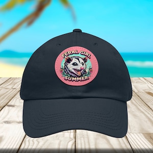 Op de afbeelding: Een marineblauwe baseballpet met een ronde roze en blauwe patch met een cartoon-opossum met de tekst "Feral Girl Summer".