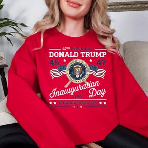 以下が含まれることがあります： 「47th President Donald Trump 45 47」のテキストと、大統領の紋章と交差したアメリカ国旗が描かれたグラフィックデザインが施された赤いスウェットシャツ。 「Inauguration Day January 20th 2025」のテキストも含まれています。
