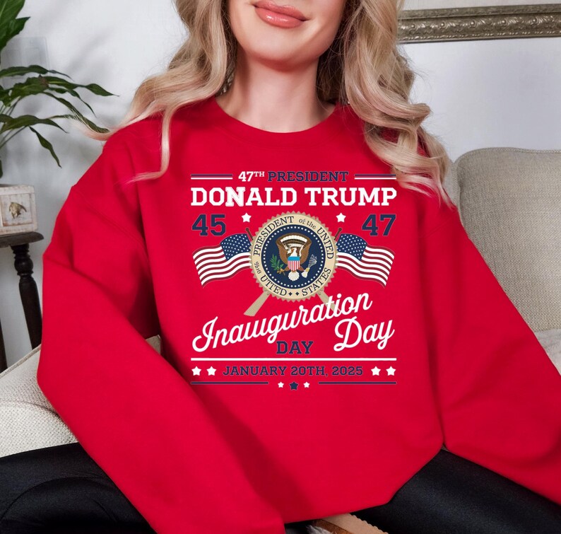 以下が含まれることがあります： 「47th President Donald Trump 45 47」のテキストと、大統領の紋章と交差したアメリカ国旗が描かれたグラフィックデザインが施された赤いスウェットシャツ。 「Inauguration Day January 20th 2025」のテキストも含まれています。