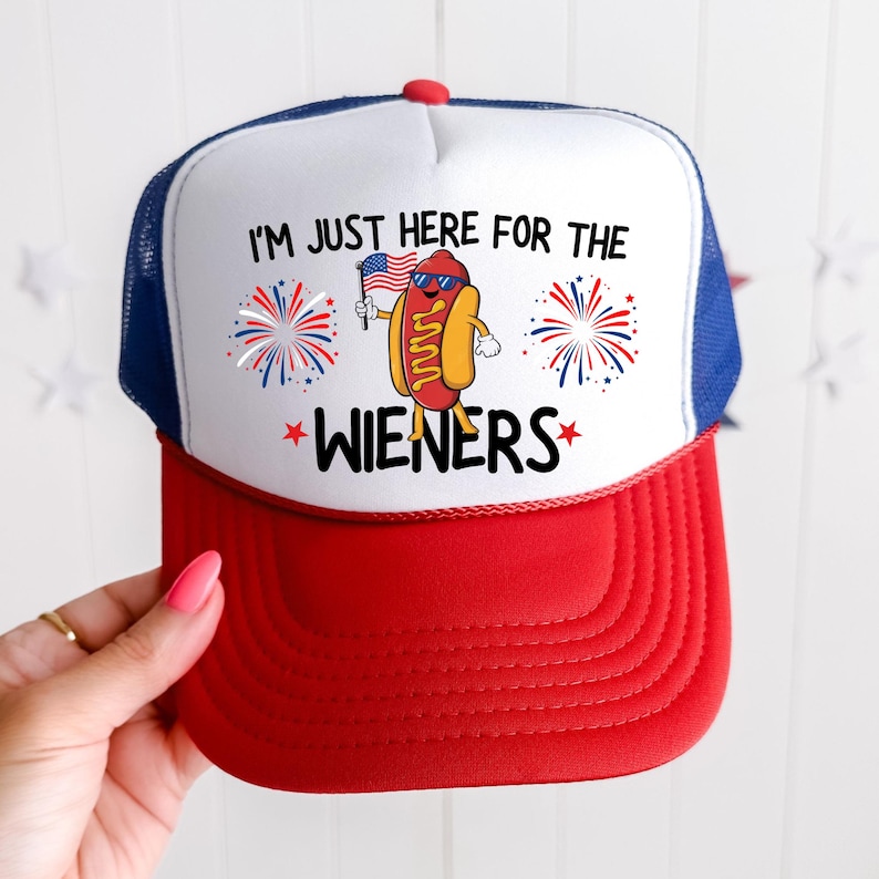 Puede incluir: Una gorra de camionero blanca y roja con una malla azul en la parte posterior. La gorra tiene un gr&aacute;fico de un hot dog sosteniendo una bandera estadounidense y fuegos artificiales con el texto "Solo estoy aqu&iacute; por las salchichas".