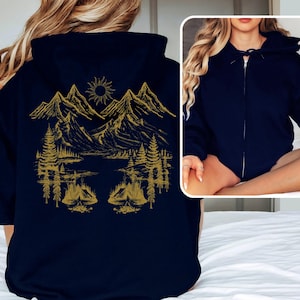 Könnte beinhalten: Ein marineblauer Reißverschluss-Hoodie mit einer goldenen Bergszene und zwei Zelten. Die Szene beinhaltet eine Sonne, Bäume und einen See.