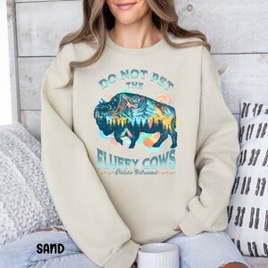 Pu&ograve; includere: Una felpa beige chiaro con una grafica di un bisonte blu e verde con montagne e alberi al suo interno. Il testo "Do Not Pet The Fluffy Cows - Nature Enthusiast -" &egrave; stampato sotto il bisonte.