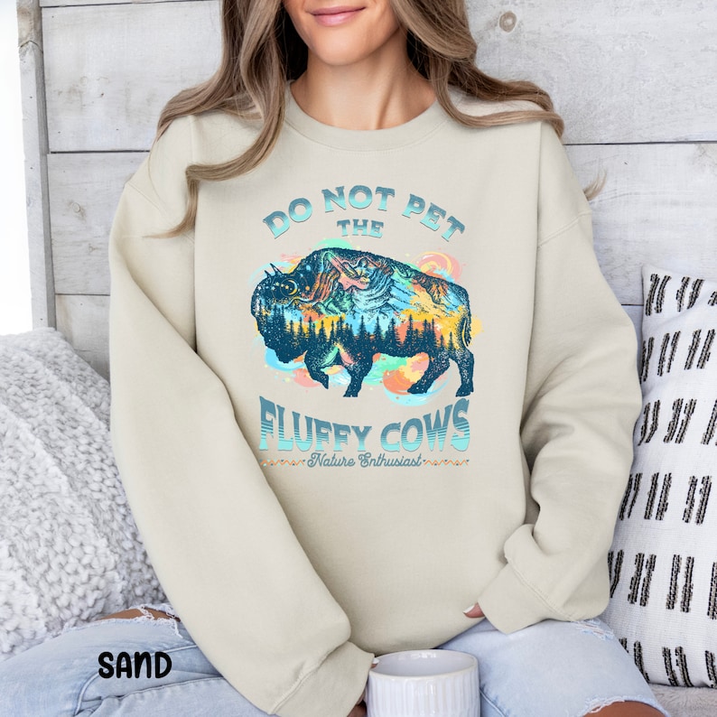 Pu&ograve; includere: Una felpa beige chiaro con una grafica di un bisonte blu e verde con montagne e alberi al suo interno. Il testo "Do Not Pet The Fluffy Cows - Nature Enthusiast -" &egrave; stampato sotto il bisonte.
