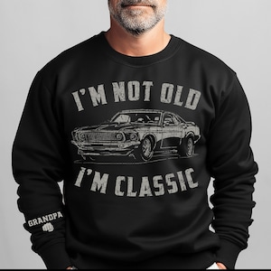 Może przedstawiać: Czarna bluza z nadrukiem w stylu vintage klasycznego samochodu i tekstem "I'm not old I'm classic". Tekst jest w stylu vintage, a samochód w stylu sylwetki. Bluza ma okrągły dekolt i długie rękawy.