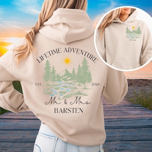 May include: A cream-coloured hoodie with a scenic design of mountains, trees, a river, and a sun. The words "LIFETIME ADVENTURE" are above the design, with "EST. 2026" and the names "Mr. & Mrs. BARSTEN" below.