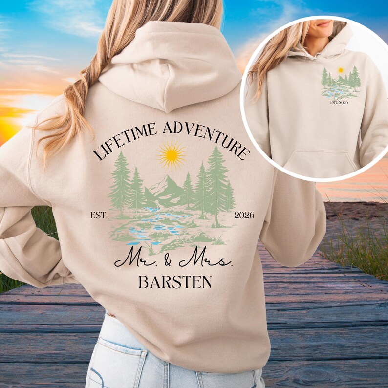 May include: A cream-coloured hoodie with a scenic design of mountains, trees, a river, and a sun. The words "LIFETIME ADVENTURE" are above the design, with "EST. 2026" and the names "Mr. & Mrs. BARSTEN" below.