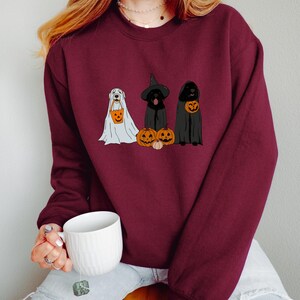 May include: Burgundy sweatshirt featuring three dogs dressed for Halloween. One dog is in a ghost costume, another in a witch hat and cloak, and the third in a cloak. Each dog holds a jack-o'-lantern bucket. A white mug is held in the person's hand.