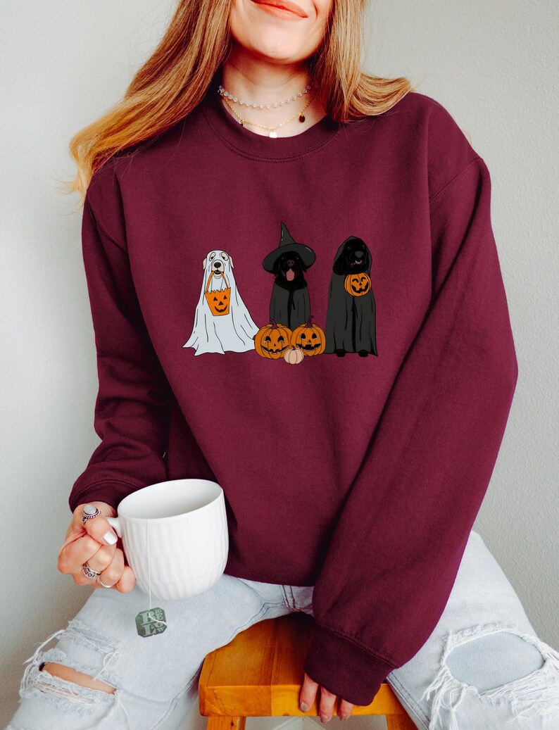 May include: Burgundy sweatshirt featuring three dogs dressed for Halloween. One dog is in a ghost costume, another in a witch hat and cloak, and the third in a cloak. Each dog holds a jack-o'-lantern bucket. A white mug is held in the person's hand.
