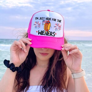 Puede incluir: Gorra de camionero rosa con una malla blanca en la parte posterior. La gorra tiene un gr&aacute;fico de un hot dog con gafas de sol y sosteniendo una bandera estadounidense con el texto "Solo estoy aqu&iacute; por las salchichas".