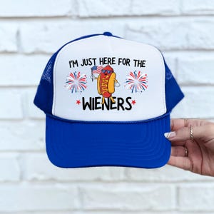 Puede incluir: Una gorra de camionero azul y blanca con una bandera estadounidense roja, blanca y azul y un perro caliente de dibujos animados que sostiene una bandera estadounidense m&aacute;s peque&ntilde;a. El texto en la gorra dice "I'm just here for the wieners".
